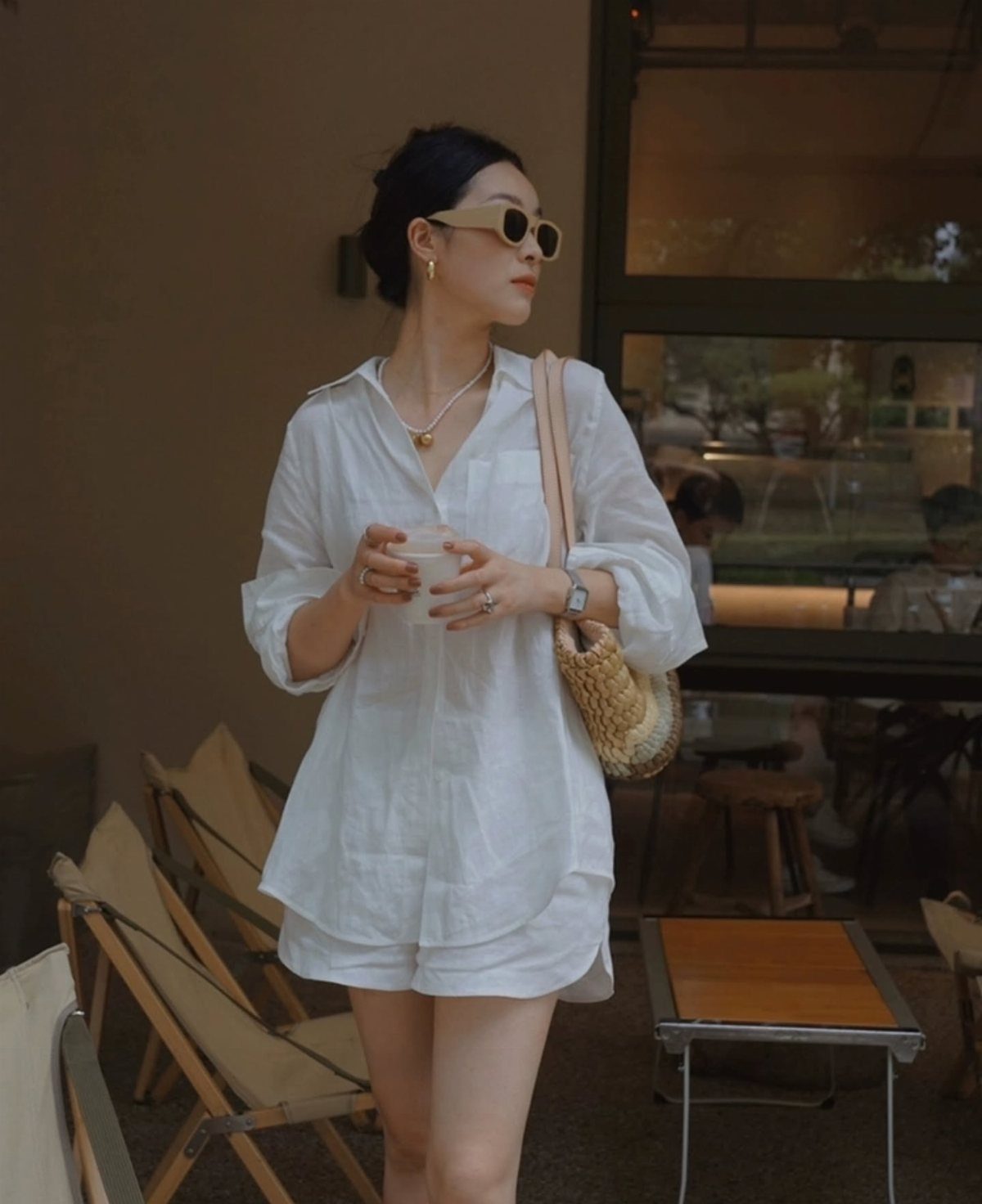Woman in oversized white shirt and white shorts holding a drink outdoors.