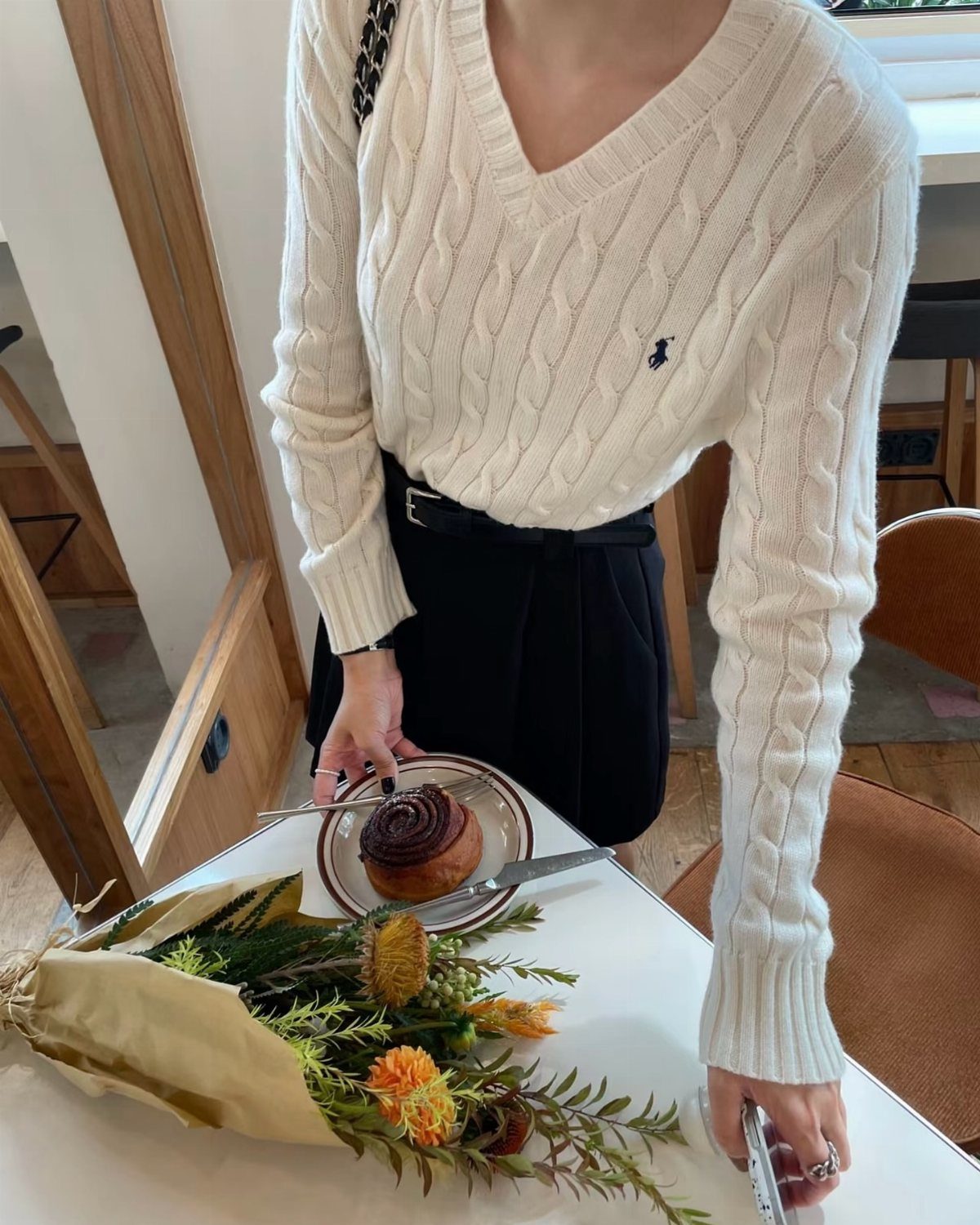 13 Winter Wear Ideas for Women: Coats, Knits and Boots 8 Woman wearing a white V-neck cable knit sweater standing by a cafe table.
