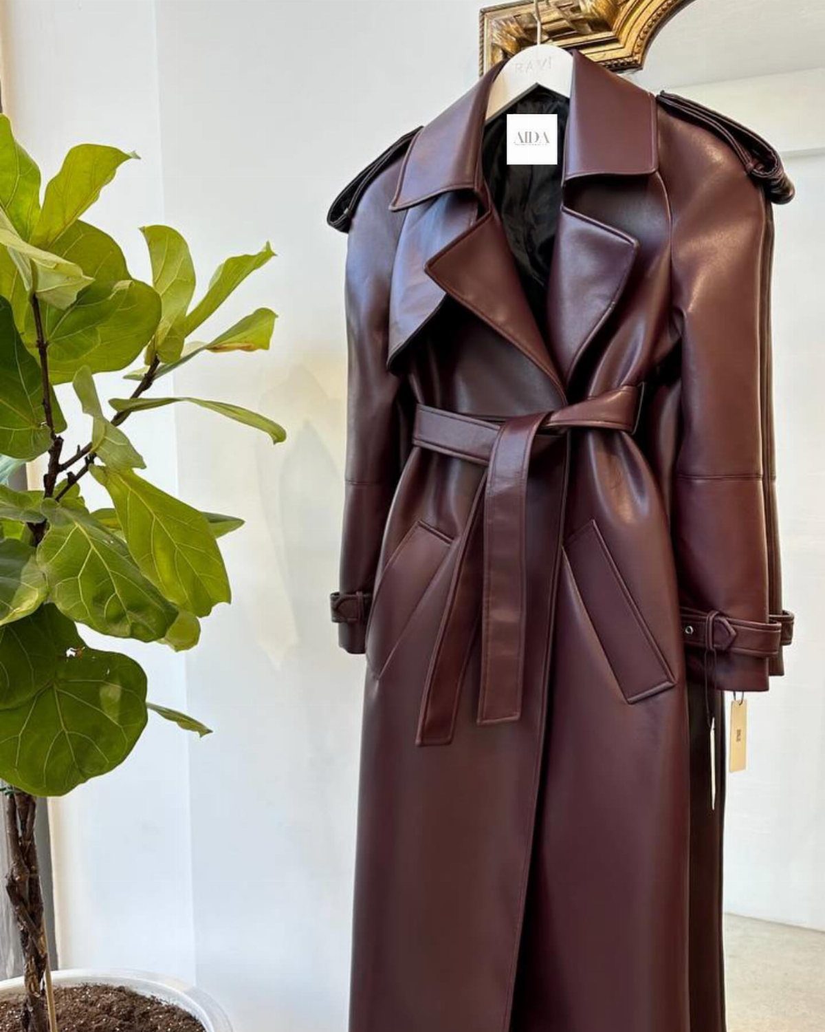13 Winter Wear Ideas for Women: Coats, Knits and Boots 3 Burgundy leather trench coat with a belt hanging next to a green plant.