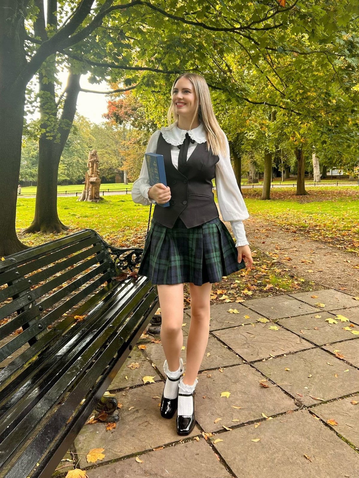 14 Women Skirt Outfits for Every Season and Occasion 14 Woman wearing a plaid pleated skirt, black vest, and white blouse in a park